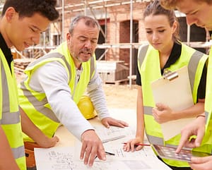 Three students being advised by a mentor on a construction site while looking at a blueprint. All four are wearing safety vests.